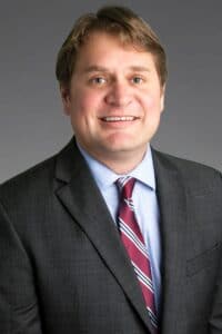 Andy Wright, a man with short, light brown hair wearing a gray suit jacket and red and white tie.